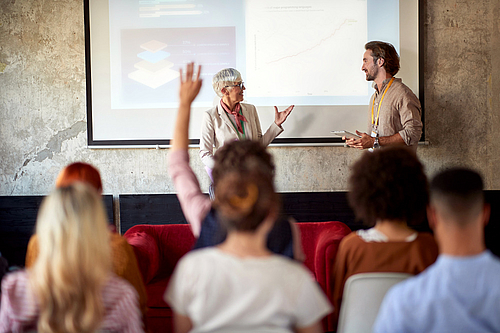 Frau hält eine Präsentation vor einer Gruppe sitzender Personen, eine Person im Publikum hebt die Hand. 
