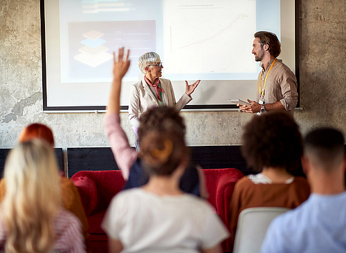 Frau hält eine Präsentation vor einer Gruppe sitzender Personen, eine Person im Publikum hebt die Hand. 