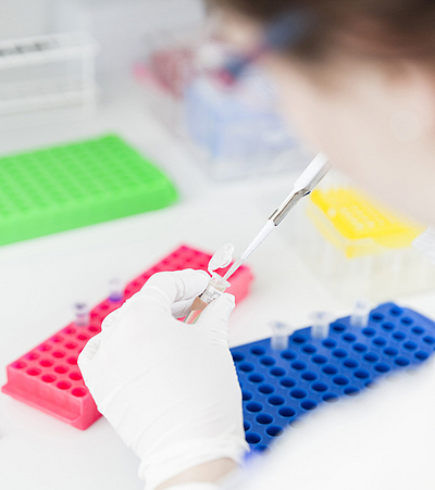 Hand mit weißem Handschuh hält eine Pipette über einem  Reaktionsgefäß vor bunten Laborständern.