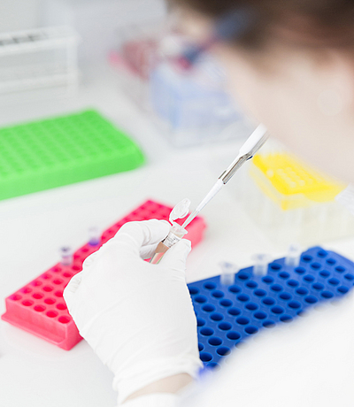Hand mit weißem Handschuh hält eine Pipette über einem  Reaktionsgefäß vor bunten Laborständern.