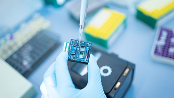 Nahaufnahme einer Hand mit Handschuh, die mit einer Pipette Flüssigkeit auf einen kleinen quadratischen Chip gibt.