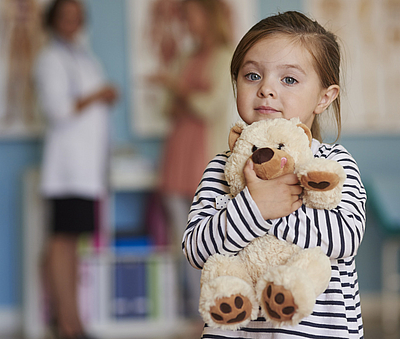 Kleines Mädchen umarmt Teddybär vor unscharfem Hintergrund mit zwei Erwachsenen. 