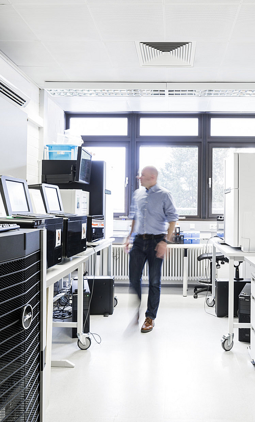 Mann geht durch hellen Laborraum mit technischen Geräten auf Tischen und großen Fenstern im Hintergrund.
